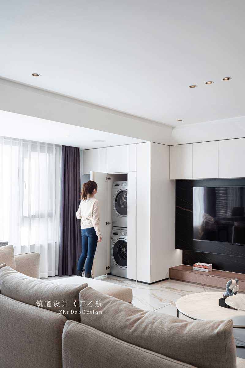 設計師充分利用陽臺的空間，把橫在客廳與陽臺的墻體打通，讓整個陽光通過大面積的窗戶照射進來，使整個空間采光通透；又將電視背景墻與之結合，并做了隱形門處理，將洗衣機、烘干機嵌入，既美觀又實用。